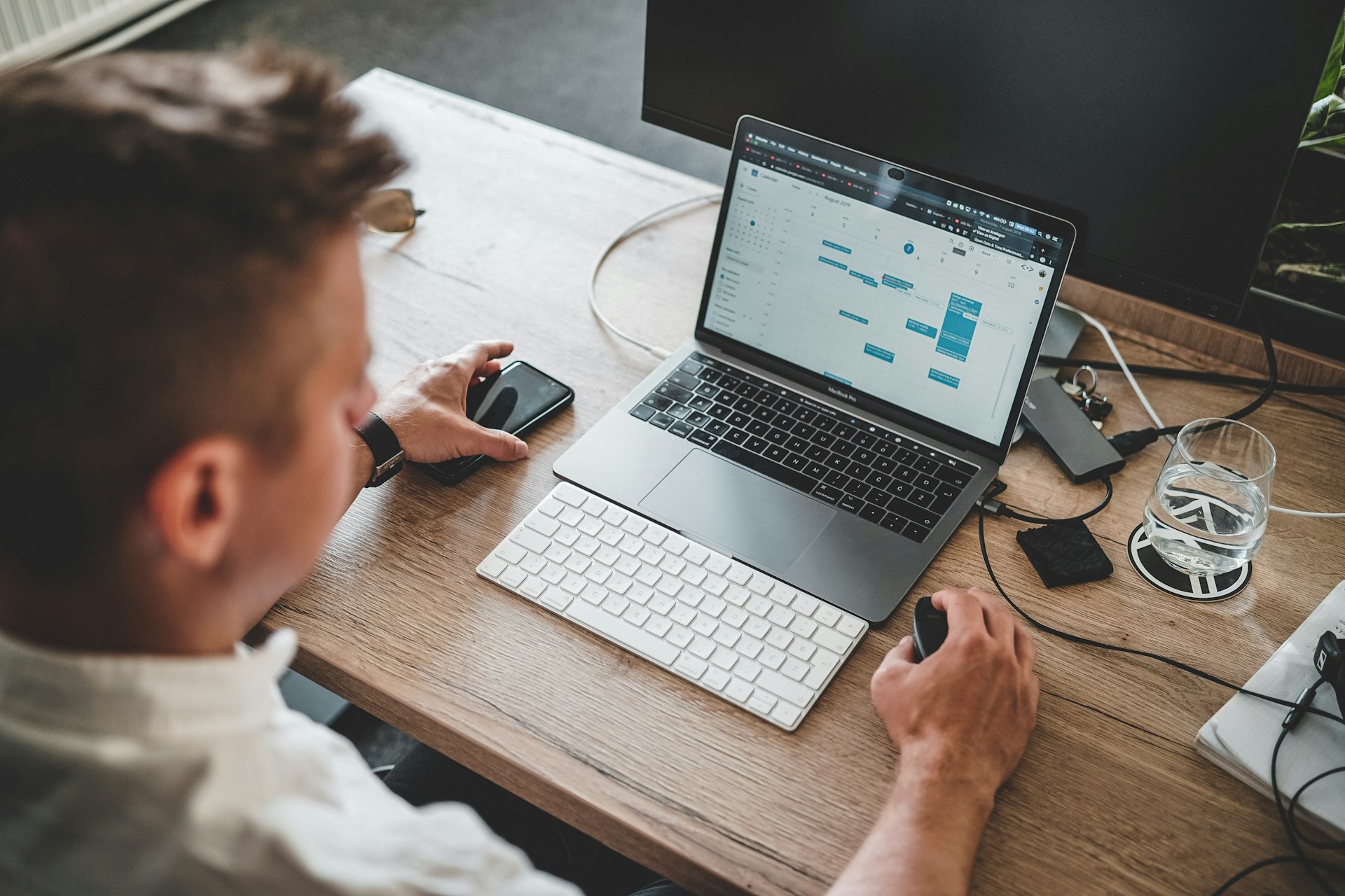 Professional Werkt op een Laptop met een Digitale Agenda Een professional werkt aan een bureau met een laptop waarop een agenda te zien is, terwijl hij een smartphone en een muis gebruikt in een moderne kantooromgeving.